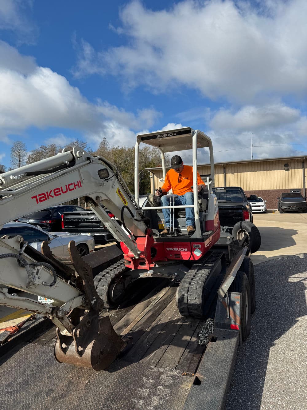 Man in orange shirt operating a Takeuchi mini excavator on a trailer at a construction site.