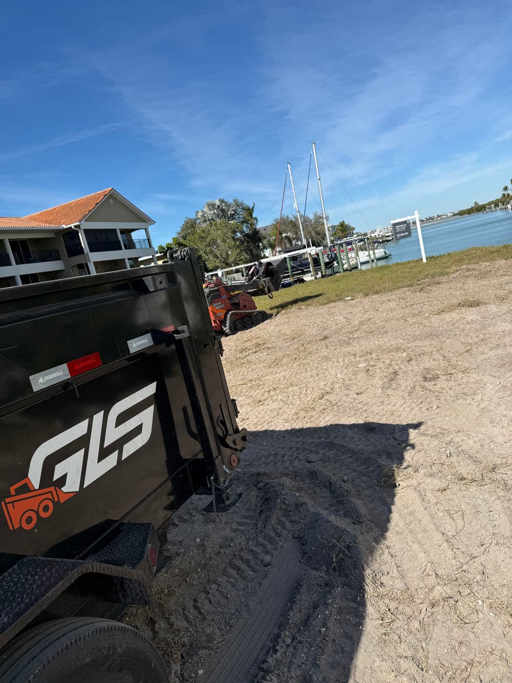 Black trailer with GLS logo on a sandy area near a marina and clear blue sky.