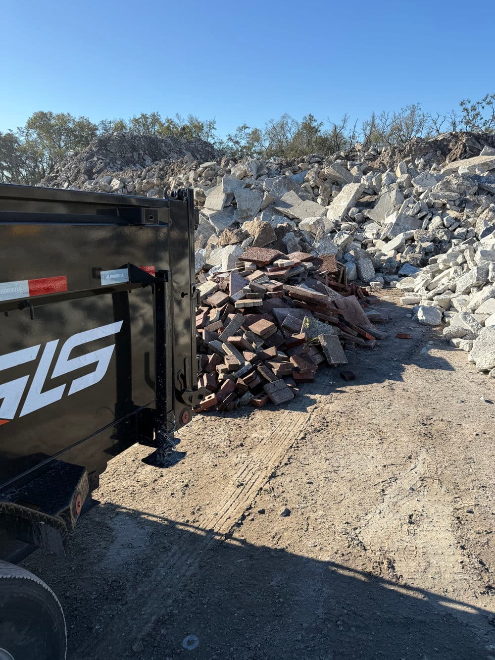 Construction debris pile beside a GLS truck at a gravel site under clear blue skies.