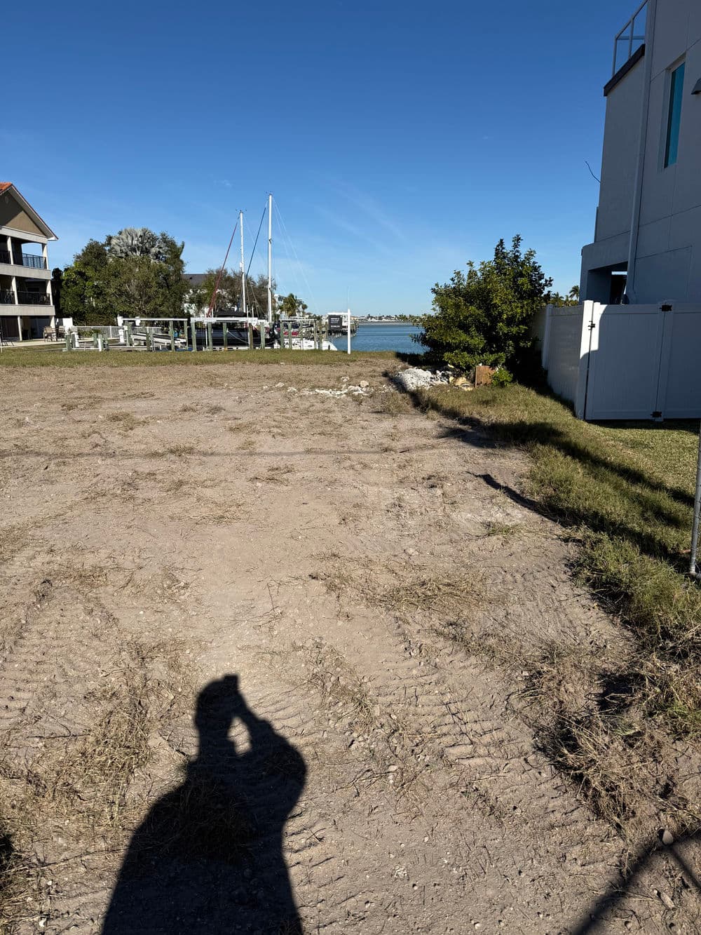 Vacant lot with grass, shadow, and marina in the background under a clear blue sky.