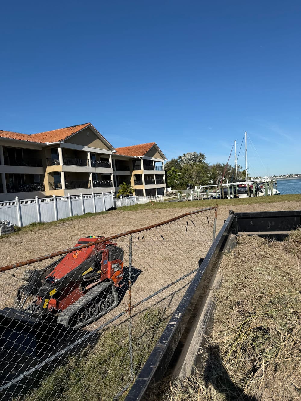 Red construction vehicle on sandy waterfront near waterfront buildings and sailboats.