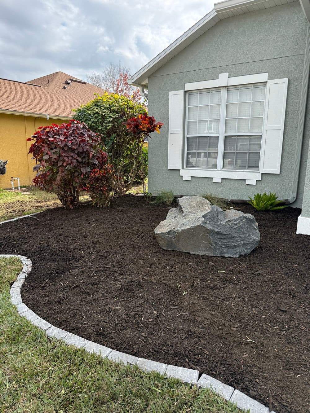 Landscaped garden with rock, mulch, shrubs, and flowers beside a gray house.