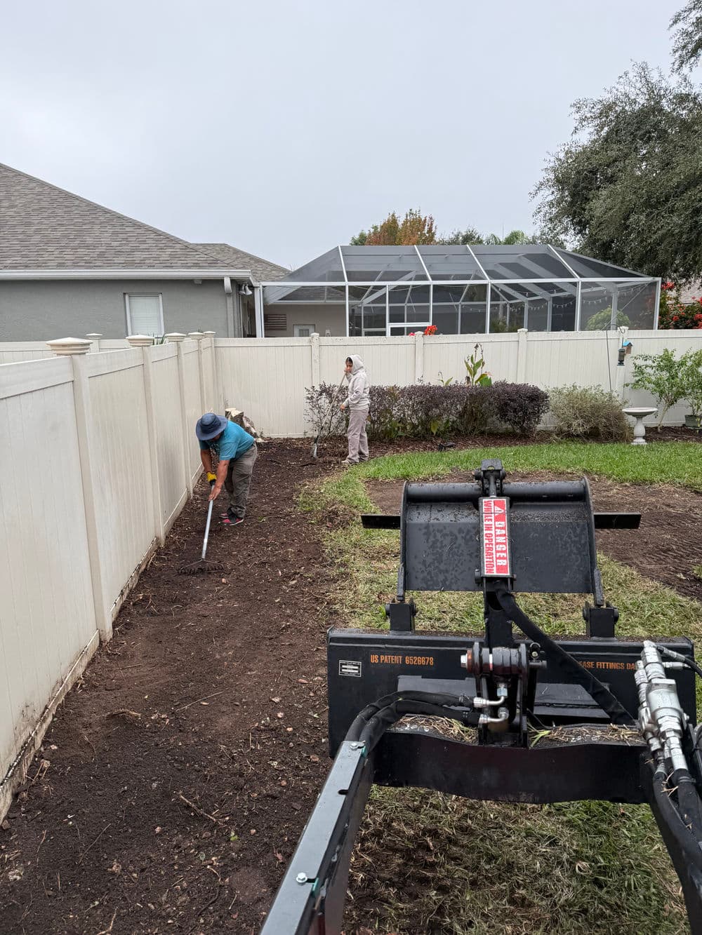 Man using a tiller in a garden while a woman plants flowers behind a fence.
