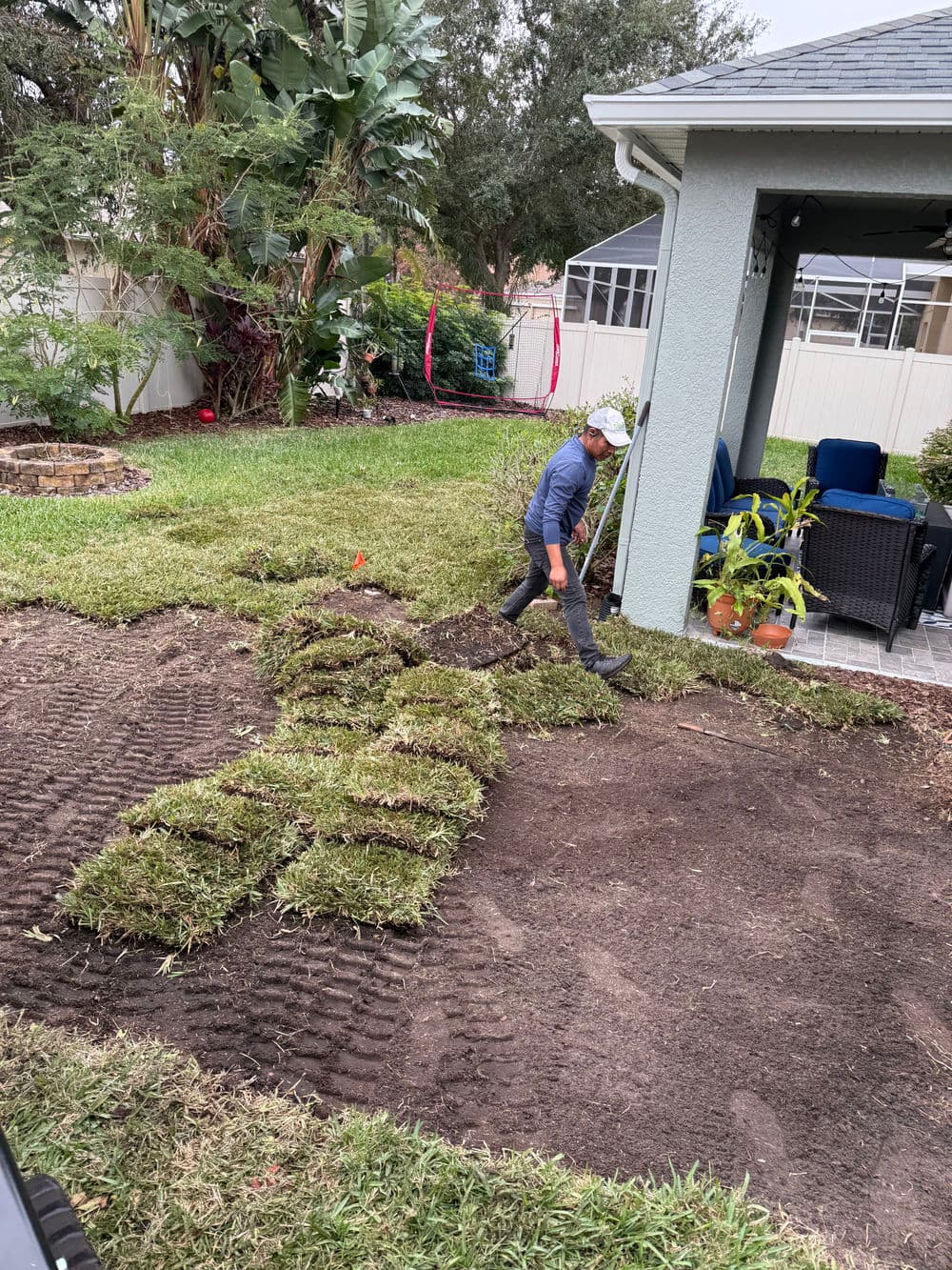 Person installing fresh sod in a residential yard, with green grass rolls and patio furniture nearby.