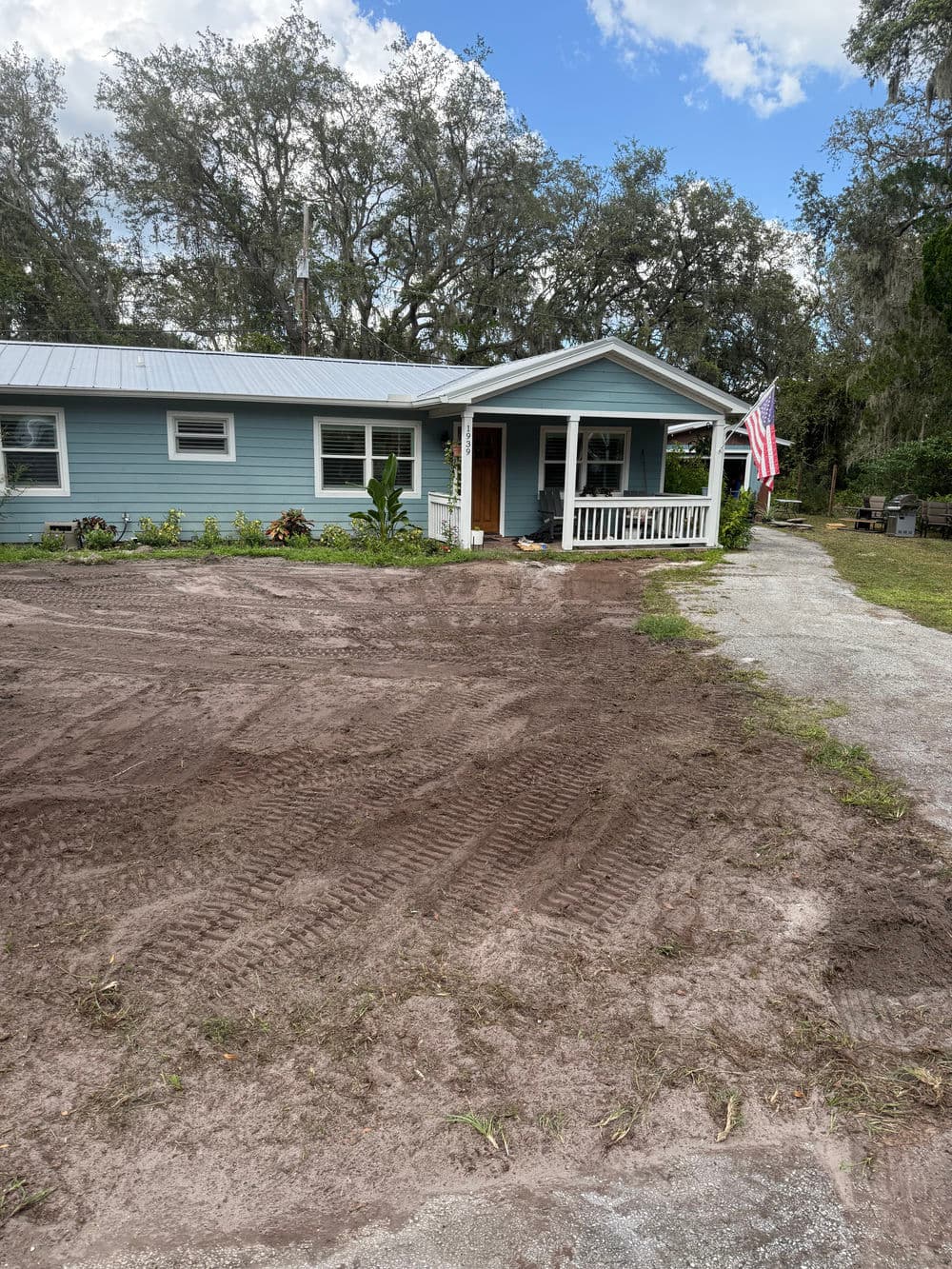 Blue house with porch and American flag, surrounded by freshly tilled soil and greenery.