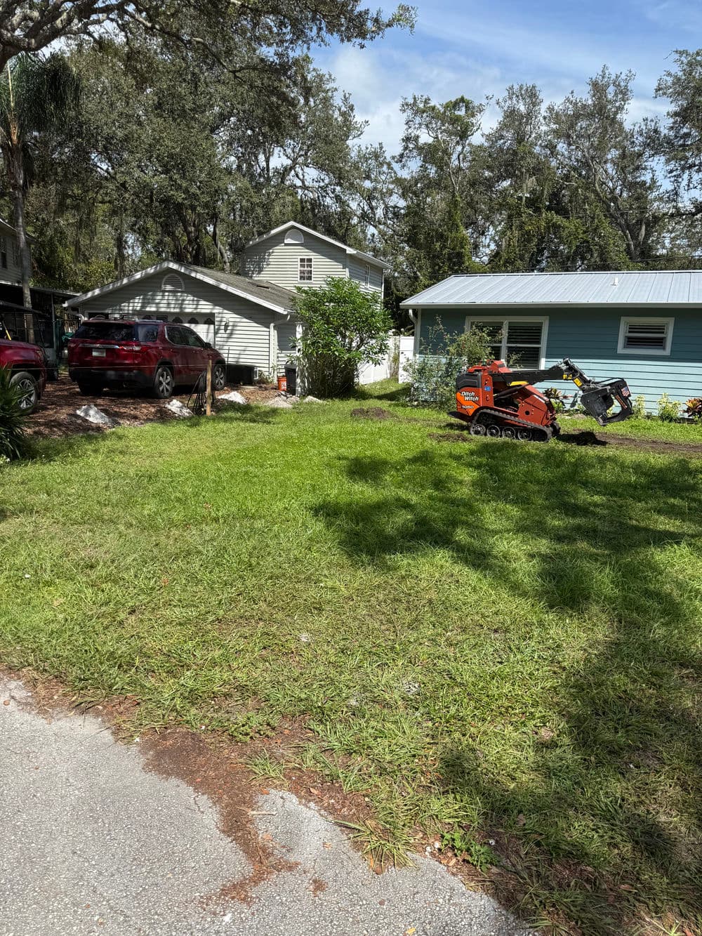 Lawn with a small landscaping tractor, two houses in background, bright sunny day.