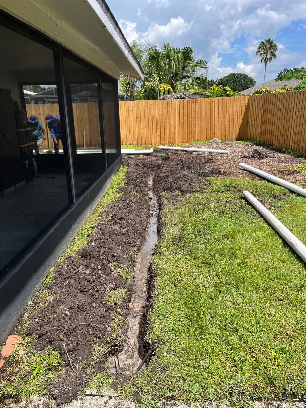 Excavated backyard trench for drainage installation beside a house with palm trees in background.