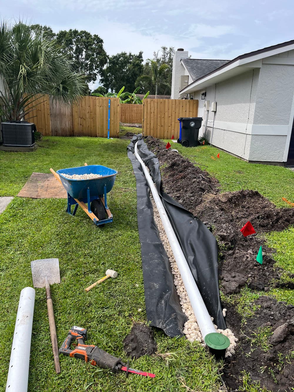 Drainage installation with a wheelbarrow, tools, and gravel in a residential yard.
