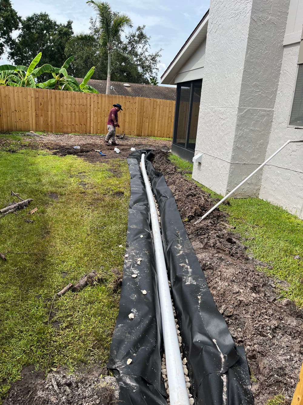 Worker installing drainage pipe in backyard with freshly dug trench and landscaping.