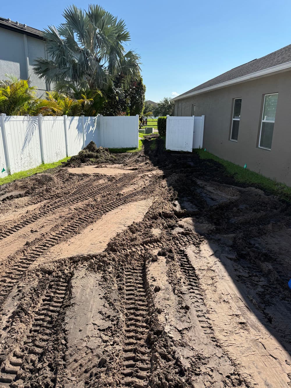 Mud-covered pathway with tire tracks alongside a house and white fencing under clear blue sky.