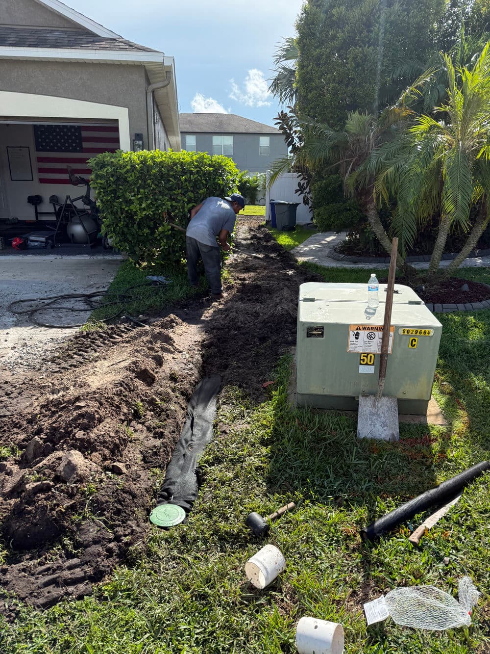 Worker digging a trench for utility installation in a residential yard with an American flag backdrop.