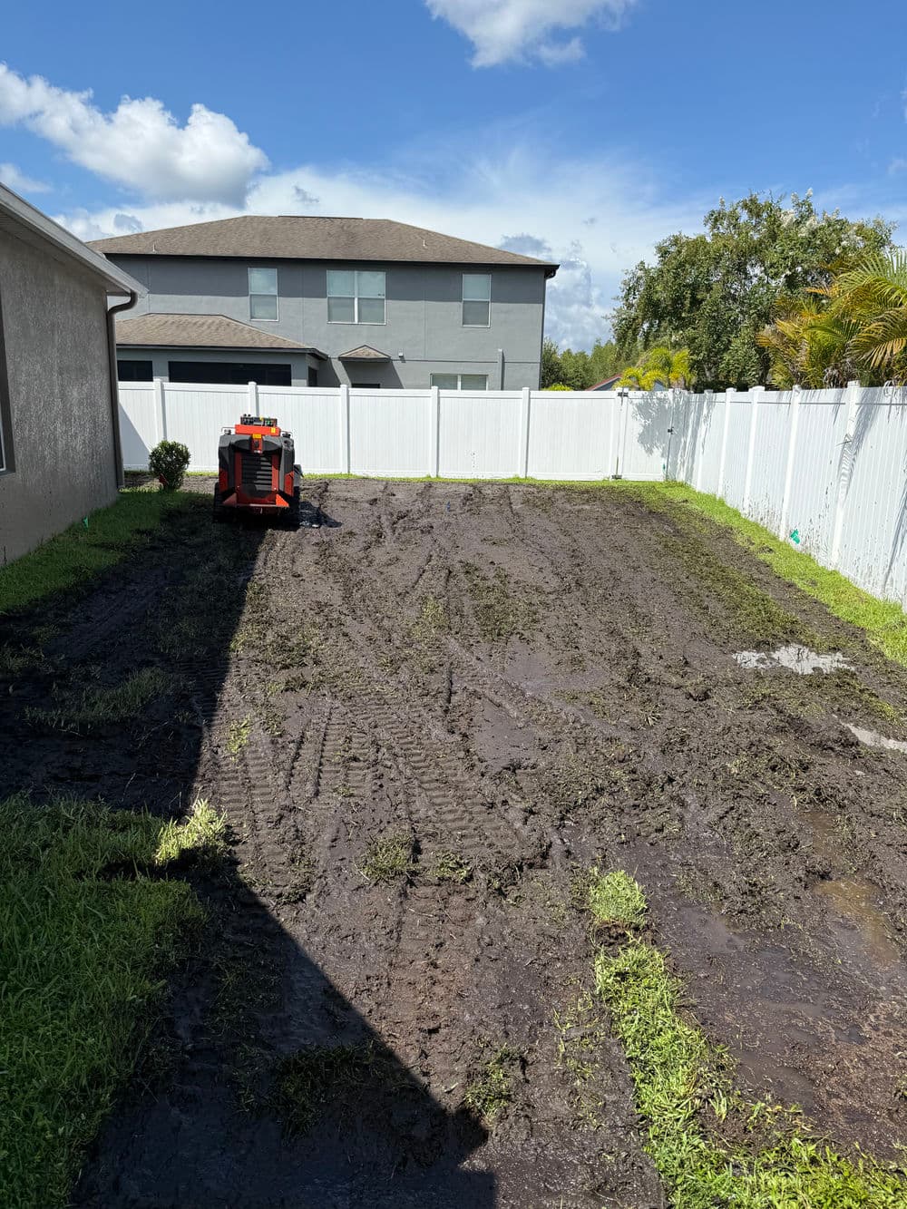 Freshly tilled muddy backyard with tractor, surrounded by white fence and blue sky.