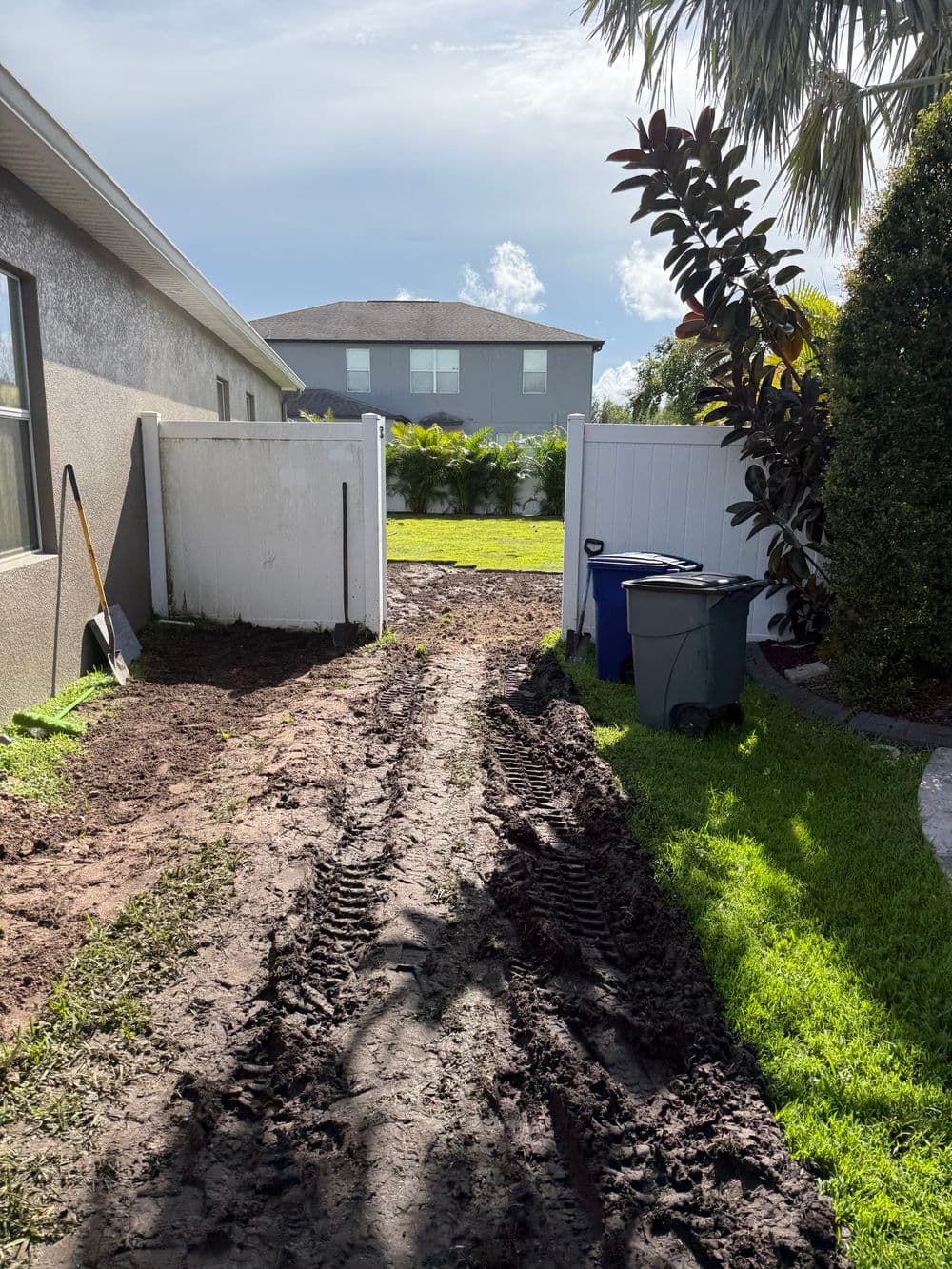 Mud path leading to a backyard with a view of a house and greenery beyond a white fence.