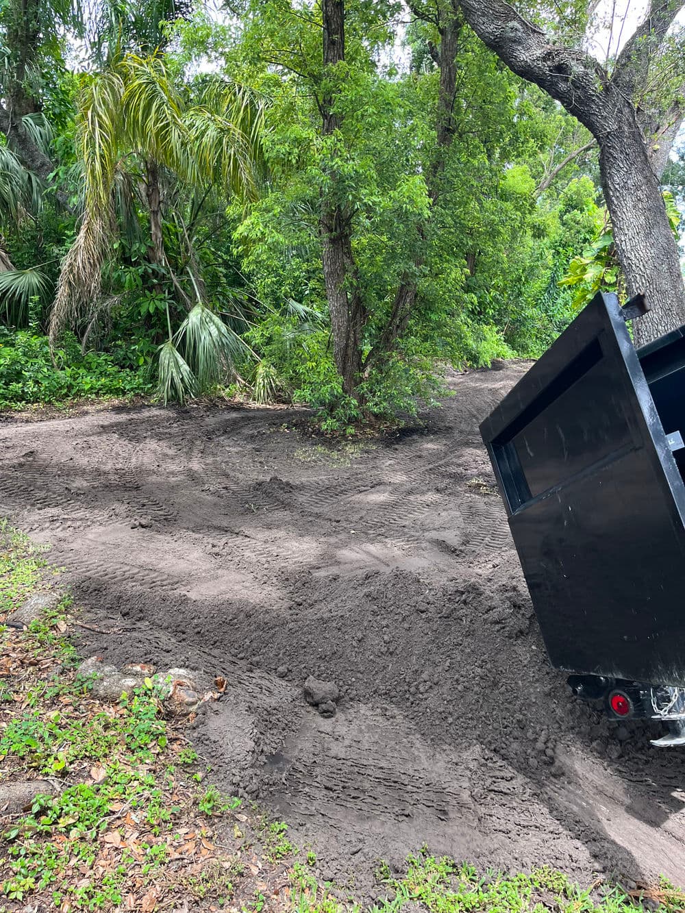 Dump truck parked on dirt riverbank surrounded by lush greenery and trees.
