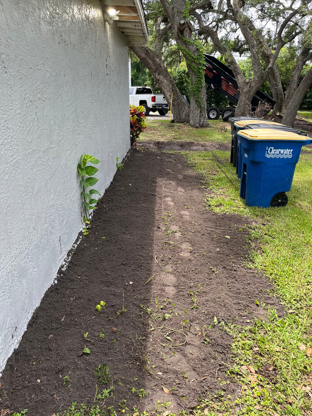 Cleared dirt area beside house with trash bins and truck in the background, ready for landscaping.