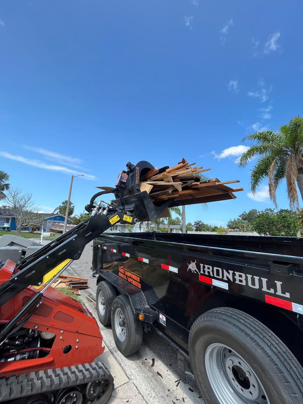 Bobcat loading lumber into an Ironbull trailer under a clear blue sky.