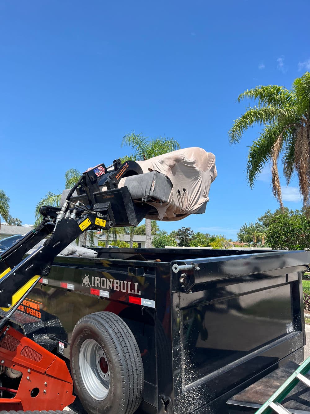 Dump truck with covered equipment against a clear blue sky and palm trees in the background.