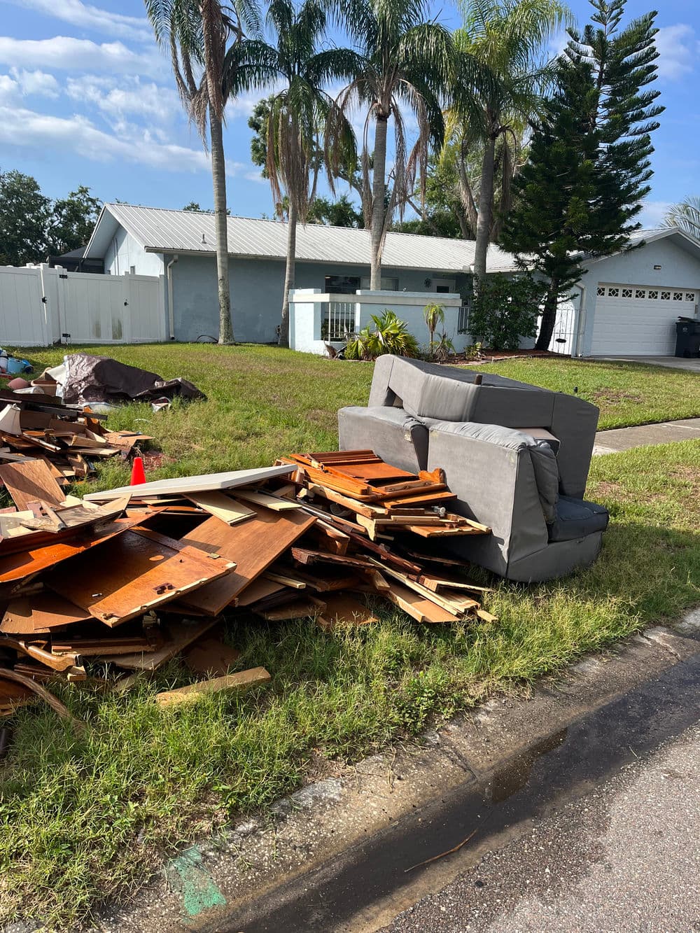 Abandoned couch and wooden debris piled on grass outside a house with palm trees.