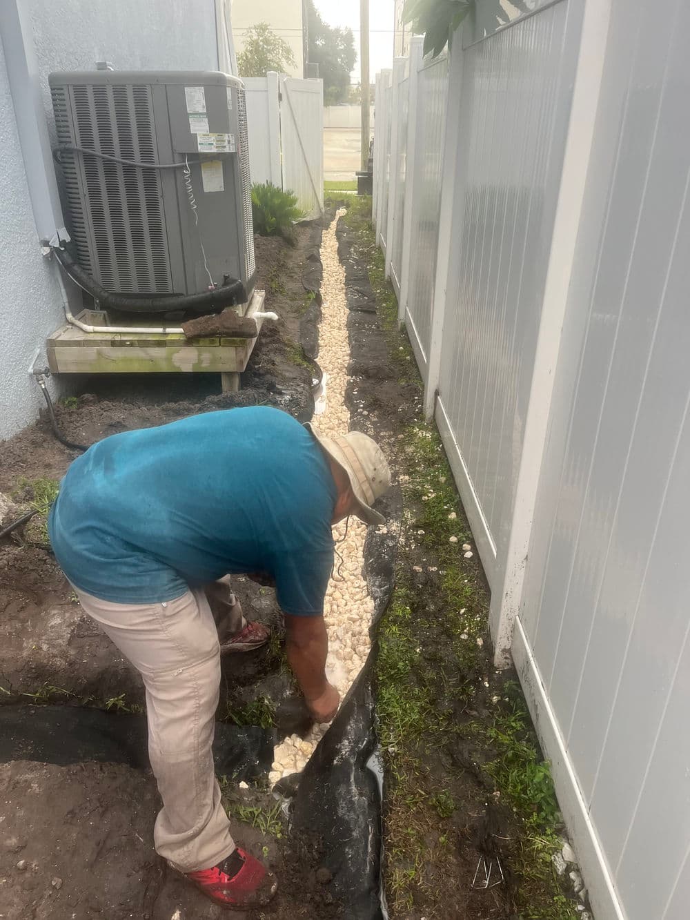 Person installing drainage gravel along a narrow trench beside a building.