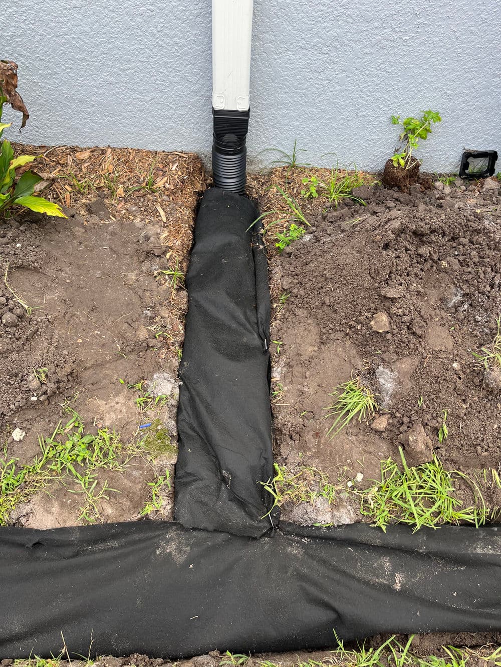 Drainage system installation with black landscape fabric and soil around downspout area.