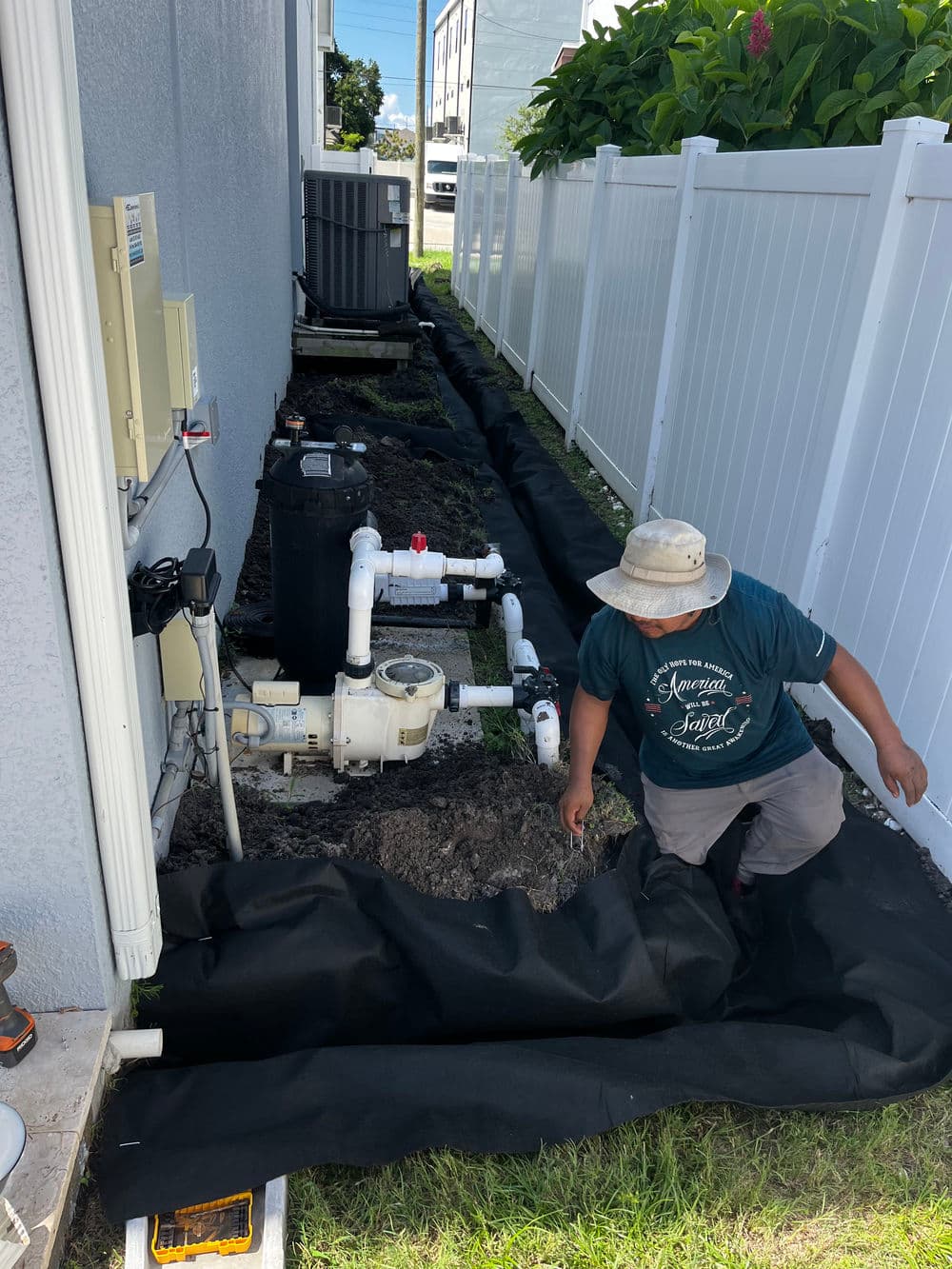 Worker installing a drainage system alongside a house, with tools and equipment visible.