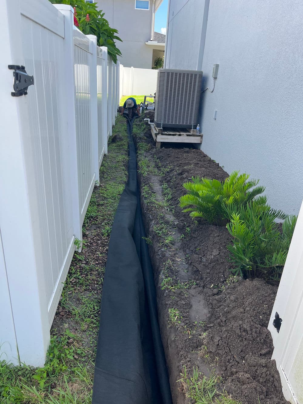 Trench installation for drainage alongside a house, with a white fence and air conditioning unit.